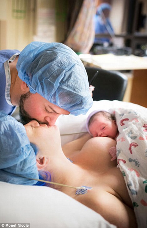 A mother kisses her partner while their baby breastfeeds in a tender family portrait despite the hospital caps and gowns, taken by Monet Nicole from Denver, Colorado