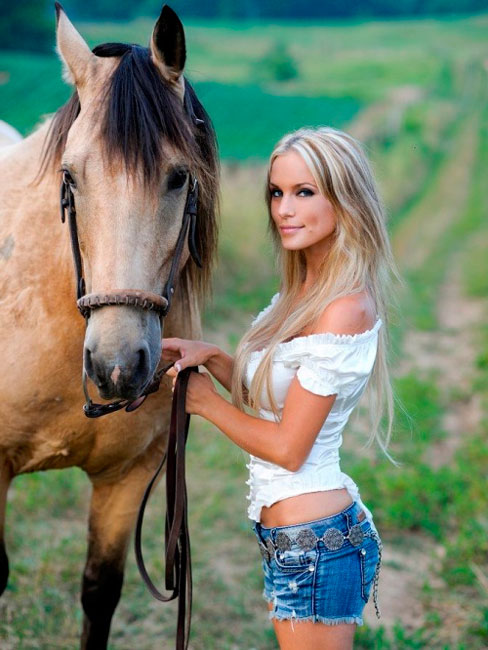 Девушка в белой блузе с открытими плечами и джинсовых шортиках, стоит рядом с лошадью